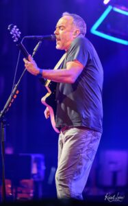 Male guitarist singing into a microphone on a stage lit with blue and purple lights.