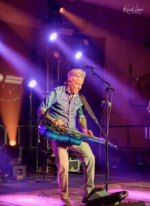 Musician with blond hair in a denim shirt playing a blue pedal steel guitar on a dark stage with purple lights.