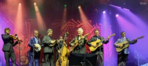 Seven musicians in suits perform on a colorful stage with violin, banjo, upright bass and guitars under vivid lights.