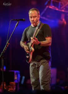 Male guitarist performing on stage, singing into a microphone while playing an electric guitar under colorful stage lights.
