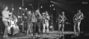 Black-and-white photo of a band performing on stage: upright bass on the left, violin, guitar, mandolin/banjo, keyboard players, and musicians under stage lights.