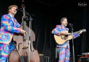 Two musicians in tie-dye suits perform on a stage; one plays a double bass and sings into a microphone while the other plays an acoustic guitar; stage gear around them.