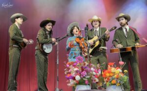 Group of five country musicians in pinstripe suits performing on stage with guitars, a banjo, and a fiddle, colorful stage lighting, and flower arrangements in front.