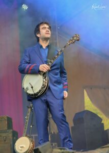 Male musician in a blue suit on stage, holding a banjo during a live performance with colorful stage lights and smoky background.