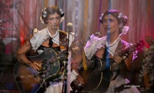 Two women in traditional white ruffled dresses playing acoustic guitars on stage, flowers in their hair with a microphone between them.