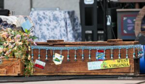 Decorated wooden float with blue fringe, tassels, and colorful souvenir license plates (West Virginia, Kentucky, Melbourne) and a bouquet of flowers on the left.