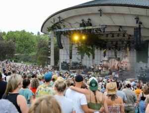 Outdoor concert scene: a large stage with bright lights and a dense crowd watching a performance, under a curved roof at Meijer Gardens with a banner overhead reading 'Fifth Third Bank Summer Concerts'.