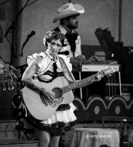 Woman in a frilly dress with a flower crown plays an acoustic guitar on stage, with a man in a cowboy hat in the background.