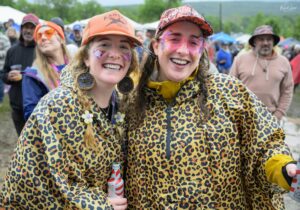 Two women in leopard-print outfits smiling at an outdoor festival, wearing face paint and hats, holding striped bottles. (informative)