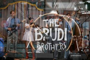A four-member live band performing on a decorated stage, with guitarist, violinist, singer, and upright bass player.