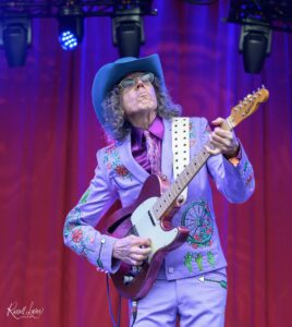 Guitarist in a lavender embroidered suit and blue hat playing an electric guitar on a purple-lit stage with sunglasses.
