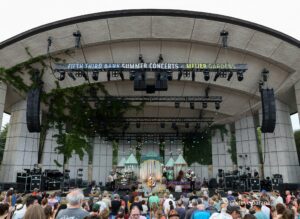 Outdoor concert stage at Meijer Gardens with a live band performing and a crowd watching the show beneath a curved roof. Banner reads 'Fifth Third Bank Summer Concerts at Meijer Gardens.'