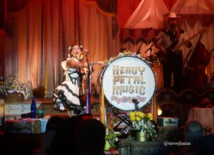 Female singer in a frilly dress sings into a microphone on a decorated stage, with a bassist and drummer in the background; front drum reads 'Heavy Metal Music'.