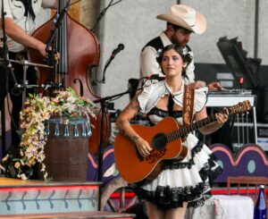 Female musician in a white frilly dress plays an acoustic guitar on stage, with flowers and band members in the background.