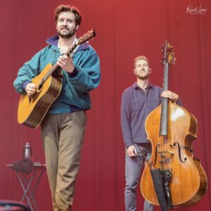 Two male musicians performing on a red-stage backdrop; one plays an acoustic guitar, the other holds a double bass beside him.