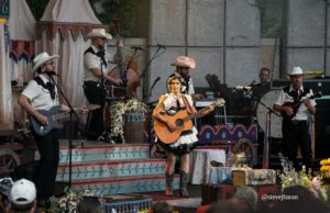 Female guitarist in a white dress with black trim and flowers in her hair performs center stage for a country band, surrounded by other musicians in cowboy hats.