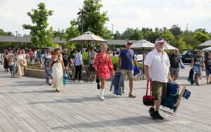 Crowd of people walking along a sunny boardwalk with trees, umbrellas, and rolling luggage, many dressed in summer clothes.