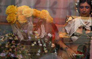 Yellow rose bouquet on display in a shop window, with a woman in a vintage outfit visible on the right behind glass.