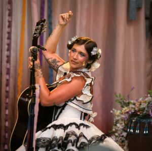 Female performer on stage raises her fist while playing a guitar, wearing a white lace dress and floral hair accessories.
