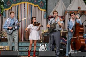 Four-member band performing live on a decorated outdoor stage with guitars, violin, and upright bass, amid colorful streamers and floral decor.