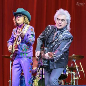 Two guitarists performing on a stage with a red curtain backdrop; one in a vibrant purple suit and turquoise hat, the other in a dark outfit with white hair playing a guitar.