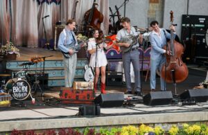 Band performing on an outdoor stage with guitars, violin, and a double bass; four musicians in vintage-style outfits.