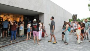People stand in a queue at an outdoor merch stall under a gray stone building, many in summer outfits and hats, some holding drinks or bags.
