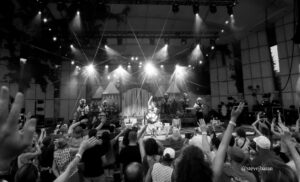 Crowd with raised hands watching a live band on a stage with bright spotlights in a large venue (black and white).