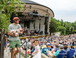 Man in a baseball cap holds a swaddled baby at a railing in the foreground, with a large outdoor concert stage and audience in the background.