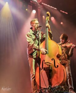 Male musician with glasses sings into a microphone while playing an upright bass on a brightly lit stage; a violinist is visible in the background.