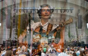 Outdoor concert scene with a band performing on a large stage; banner reads 'Fifth Third Bank Summer Concerts at Meijer Gardens' and a crowd fills the foreground.