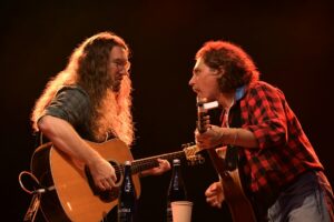 Two guitarists perform on stage: one with long hair and glasses, the other in a red plaid shirt, under warm red lighting.