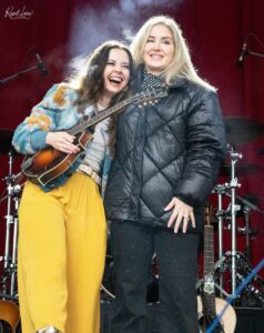 Two smiling women on a stage posing together; one holds a guitar and wears a floral blue jacket and yellow pants, the other in a black puffer jacket with black pants, drums in the background.