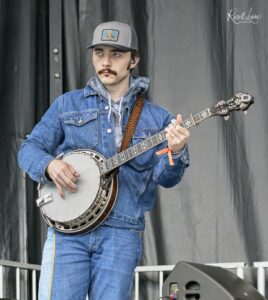 Male musician playing a banjo on stage, wearing a denim jacket and gray cap with a logo, focused on his performance