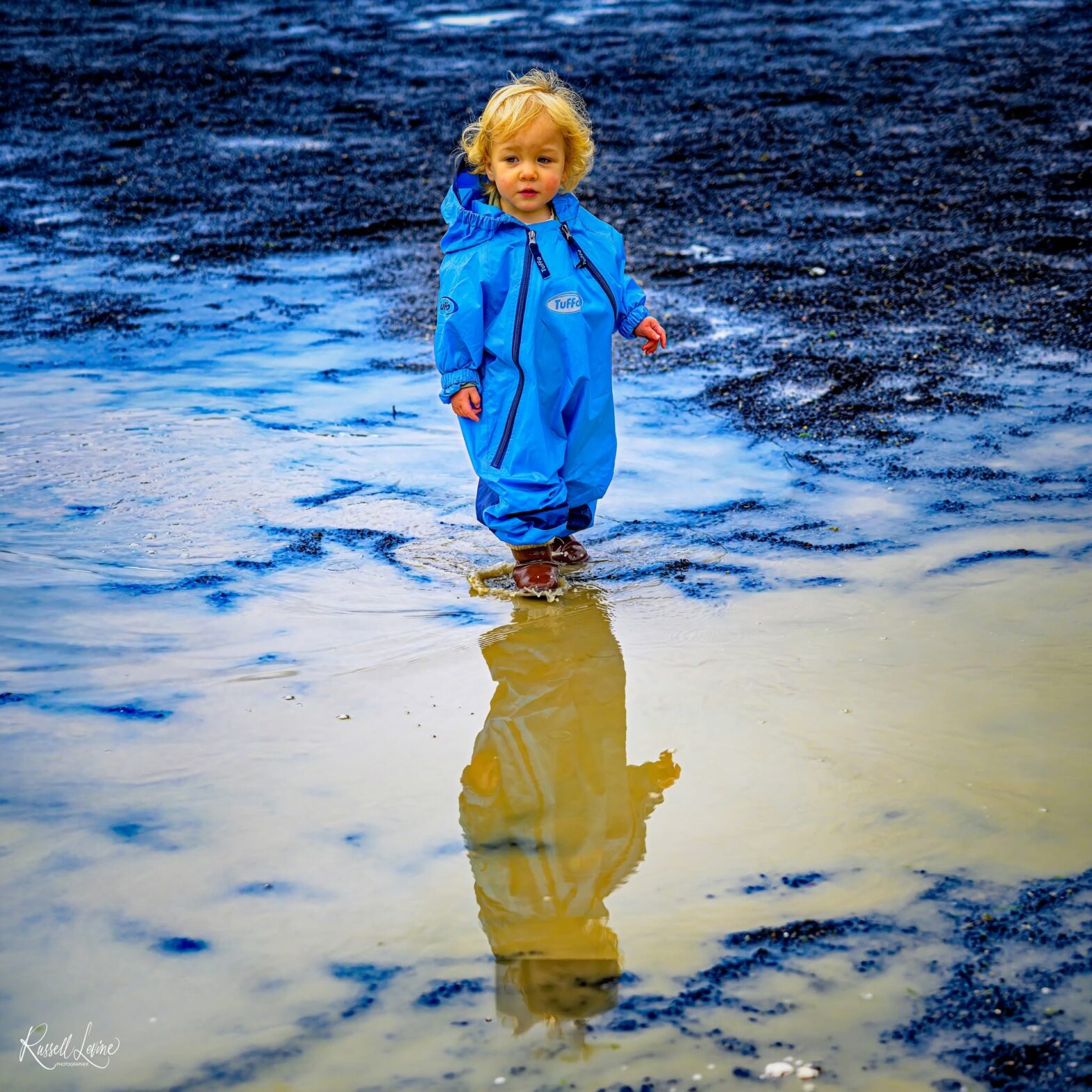 Child in a bright blue waterproof suit stands in shallow water on a rocky beach, reflection visible in a puddle beneath them.