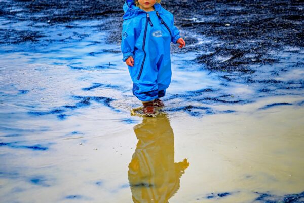 Child in a bright blue waterproof suit stands in shallow water on a rocky beach, reflection visible in a puddle beneath them.
