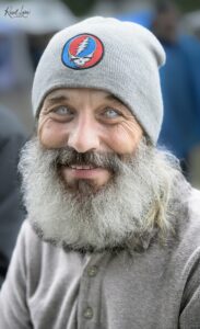 Close-up of a smiling man with blue eyes wearing a gray knit beanie featuring a red-blue Grateful Dead skull patch.