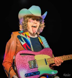 Energetic guitarist in a bright patchwork jacket and rainbow hat, playing a glittery pink guitar on a dark stage.