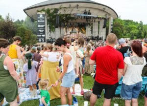 Crowd enjoying an outdoor festival in a park, facing a large open-air stage with musicians.