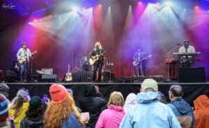 Band performs on a brightly lit stage with colorful spotlights; center singer with acoustic guitar, others on guitar, bass, drums, and keys; audience in rain ponchos in foreground.