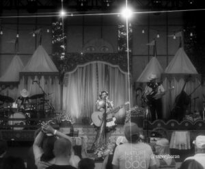 Female performer with guitar singing on an outdoor stage, decorative tents flanking the stage and string lights above, audience in foreground (black-and-white photo).