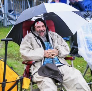 Bearded man in a beige raincoat seated in a wheelchair, laughing under a large umbrella on a rainy day outdoors.