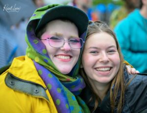 Two friends smiling for a close-up selfie, one wearing a bright green and purple scarf and a yellow jacket.