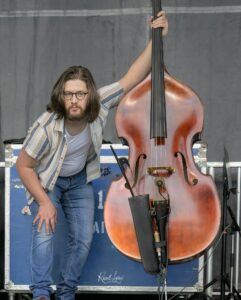 Musician with glasses and shoulder-length hair leaning on a double bass on stage during a performance.
