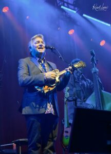 Male musician in a gray suit singing into a microphone while playing a mandolin on stage under blue lighting