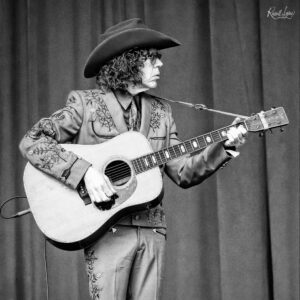 Musician wearing a wide-brim hat and glasses, playing an acoustic guitar on stage in a black-and-white photo with embroidered clothing.