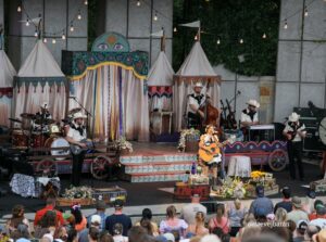 Live band performing on a decorated outdoor stage with string lights, audience watching a country festival.