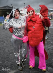 Two people in costumes pose together at an outdoor event: one in a shiny silver spacesuit and the other in a bright red lobster outfit, smiling with arms around each other.