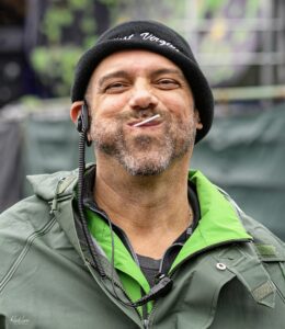 Close-up of a smiling man wearing a green outdoor jacket and black beanie, with a headset and a cigarette in his mouth.