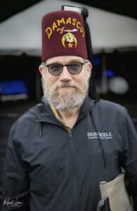 Portrait of a man with a gray beard and glasses wearing a maroon fez with Damascus insignia and dagger, outdoors in a dark jacket.